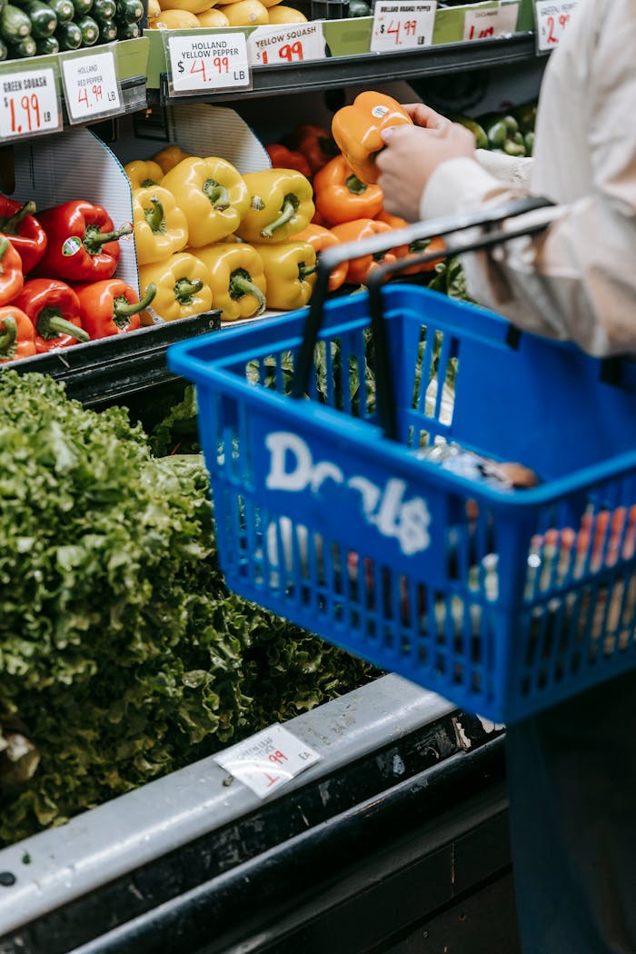 Contacto Side view of crop faceless buyer in casual clothes carrying shopping basket while choosing assorted colorful bell peppers in supermarket